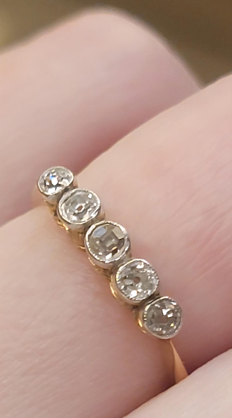 May include: A gold ring featuring five round, clear, faceted stones set in a row. Each stone is held in a circular metal setting, creating a continuous line of sparkle across the band. The design highlights the clarity and brilliance of the stones.