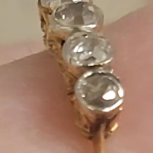 May include: A gold ring featuring a row of four round, clear, faceted stones set in silver-toned metal settings. The band is thin and gold-colored, with the stones arranged vertically along the top of the ring, showcasing a reflective, polished finish.