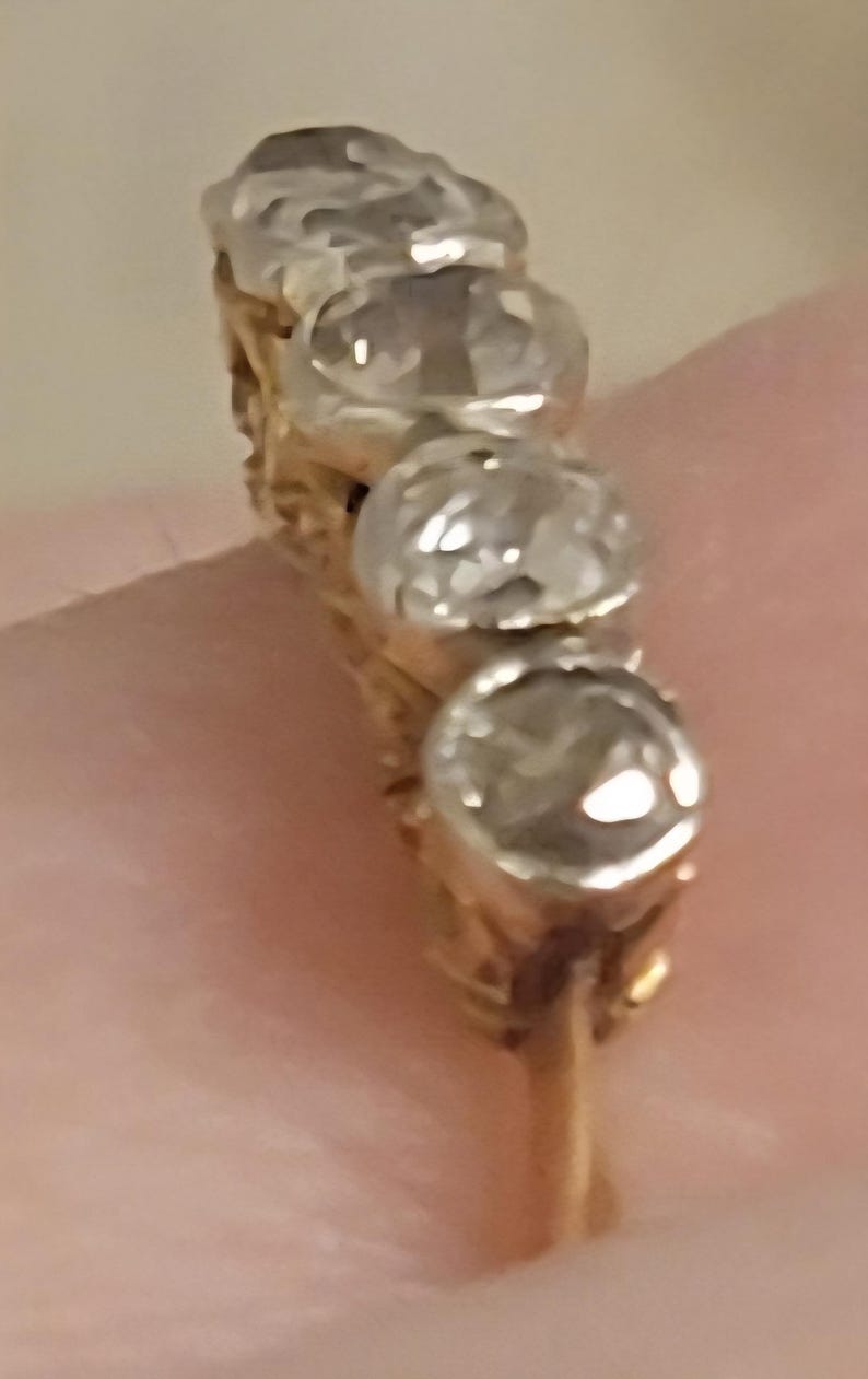 May include: A gold ring featuring a row of four round, clear, faceted stones set in silver-toned metal settings. The band is thin and gold-colored, with the stones arranged vertically along the top of the ring, showcasing a reflective, polished finish.