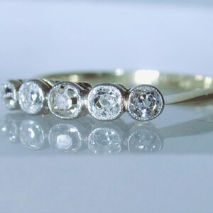 May include: A gold ring featuring five round, clear, faceted stones set in a row. Each stone is secured within a circular metal bezel setting. The band is thin and polished, reflecting light across its surface, creating a clear, bright appearance.