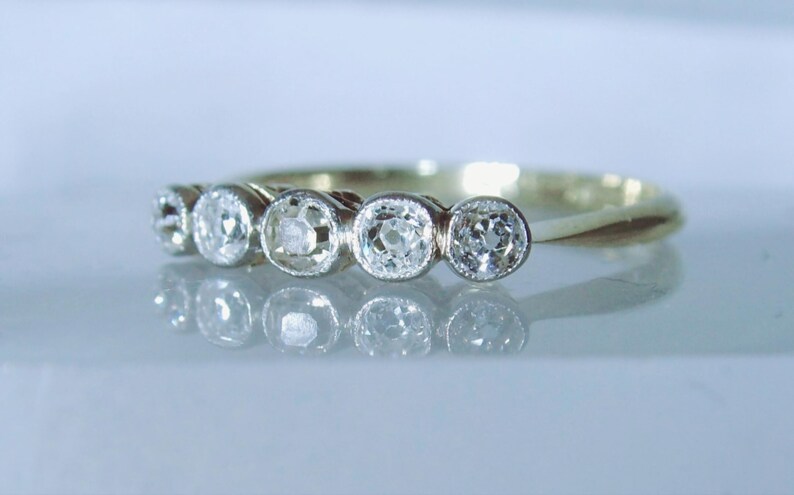 May include: A gold ring featuring five round, clear, faceted stones set in a row. Each stone is secured within a circular metal bezel setting. The band is thin and polished, reflecting light across its surface, creating a clear, bright appearance.