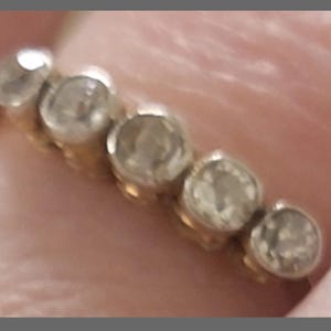 May include: A gold band ring featuring five round, clear, faceted stones set in a row. The stones are arranged horizontally across the top of the band, each held in a circular metal setting. The metal appears to be a warm yellow gold tone.