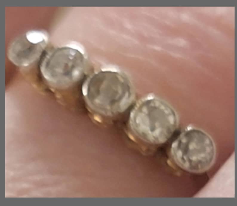 May include: A gold band ring featuring five round, clear, faceted stones set in a row. The stones are arranged horizontally across the top of the band, each held in a circular metal setting. The metal appears to be a warm yellow gold tone.