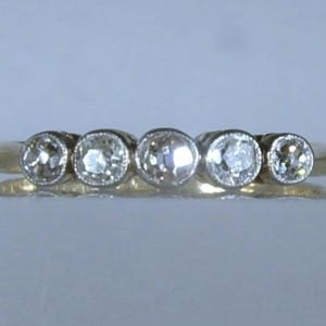 May include: A gold ring featuring five round, clear, faceted stones set in a horizontal row. Each stone is secured within a circular bezel setting, creating a continuous line of gems across the top of the polished gold band.