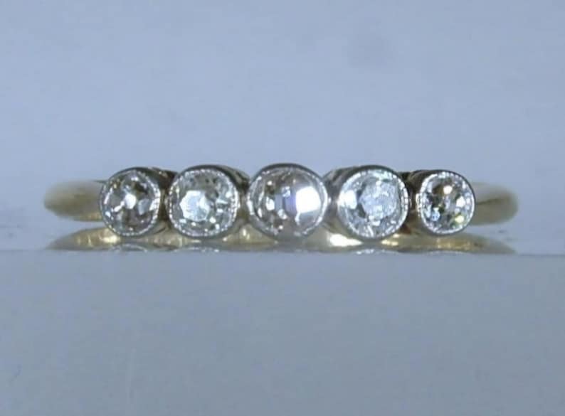 May include: A gold ring featuring five round, clear, faceted stones set in a horizontal row. Each stone is secured within a circular bezel setting, creating a continuous line of gems across the top of the polished gold band.