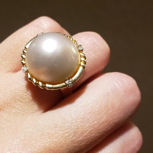 May include: A gold ring with a large, white pearl set in a rope-like design. The ring is accented with small diamonds.