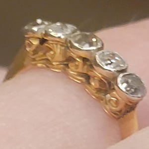 May include: A gold ring featuring five round, clear, faceted stones set in a row. The band has an intricate, textured, scroll-like metalwork design along the sides, supporting the stones in a horizontal arrangement across the top of the ring.