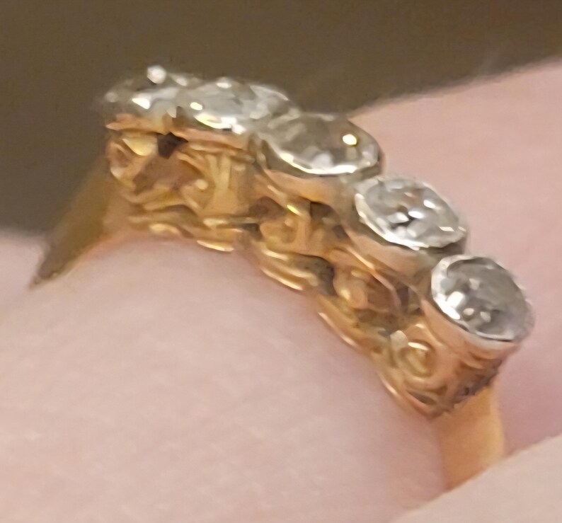 May include: A gold ring featuring five round, clear, faceted stones set in a row. The band has an intricate, textured, scroll-like metalwork design along the sides, supporting the stones in a horizontal arrangement across the top of the ring.