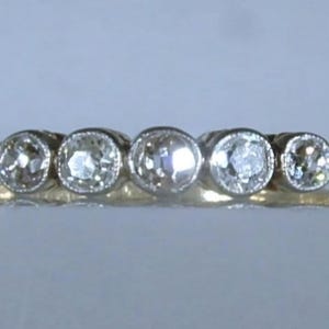 May include: A gold ring featuring five round, clear, faceted stones set in a horizontal row. Each stone is secured within a circular metal bezel setting, creating a continuous line of gems across the top of the band, which has a smooth, polished finish.