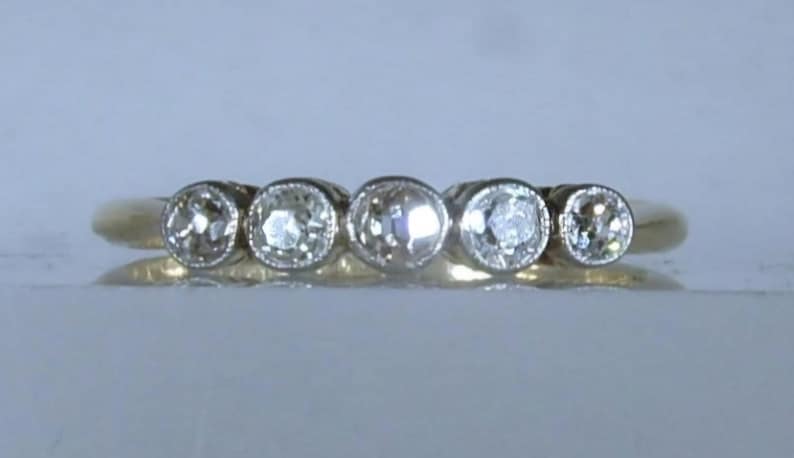 May include: A gold ring featuring five round, clear, faceted stones set in a horizontal row. Each stone is secured within a circular metal bezel setting, creating a continuous line of gems across the top of the band, which has a smooth, polished finish.