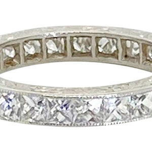 May include: A silver-toned eternity band ring featuring a continuous row of square-cut clear stones set in a metallic channel. The band displays a uniform design with visible metal prongs securing each individual stone around the entire circumference.
