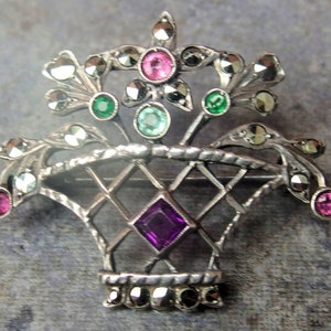 May include: A silver-toned brooch shaped like a basket of flowers, featuring a central purple square gemstone, pink and green round accent stones, and numerous small, dark, faceted marcasite stones embedded throughout the metal frame.