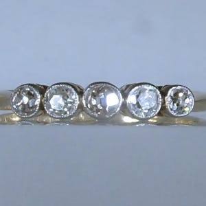 May include: A gold ring featuring five round, bezel-set clear stones arranged in a horizontal row across the top. The band is smooth and polished, with the central stone being slightly larger than the two stones on either side, creating a graduated look.