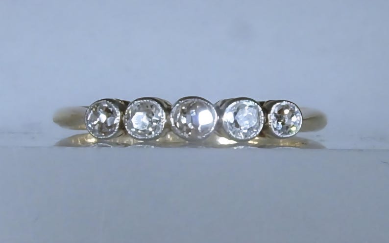 May include: A gold ring featuring five round, bezel-set clear stones arranged in a horizontal row across the top. The band is smooth and polished, with the central stone being slightly larger than the two stones on either side, creating a graduated look.