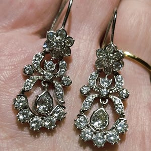 May include: A pair of silver-toned drop earrings featuring a floral-shaped top cluster and a dangling pear-shaped center stone, all encrusted with numerous small, clear, sparkling round-cut stones set in a decorative, open-work metal frame.
