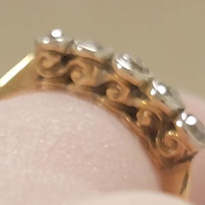 May include: A gold ring featuring a row of five clear, round-cut stones set in a silver-toned metal. The side of the ring band displays a decorative, repeating scrollwork pattern in gold, connecting the stone settings to the solid gold band.