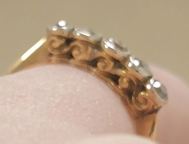 May include: A gold ring featuring a row of five clear, round-cut stones set in a silver-toned metal. The side of the ring band displays a decorative, repeating scrollwork pattern in gold, connecting the stone settings to the solid gold band.