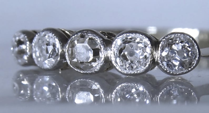 May include: A silver-toned metal ring featuring five round, faceted clear stones set in a row. Each stone is secured within a circular bezel setting with a fine milgrain edge detail along the perimeter of the metal surrounding the stones.