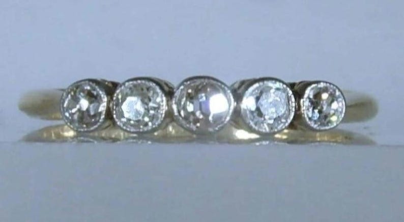 May include: A gold ring featuring five round, clear, faceted stones set in a row. Each stone is secured in a circular bezel setting with a milgrain edge. The band is smooth and polished, showcasing a consistent yellow gold tone throughout the design.