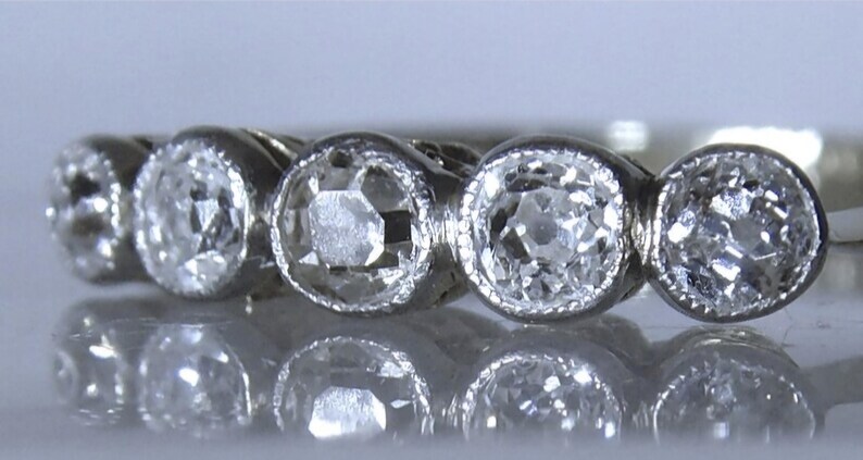 May include: A silver-toned ring featuring five round, clear, faceted stones set in a row. Each stone is secured within a circular bezel setting that has a fine, beaded texture along the outer edge, creating a continuous, elegant, and reflective band.