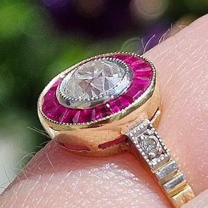 Puede incluir: Un anillo de oro con un gran diamante redondo rodeado por un halo de rubíes rojos. La banda está engastada con diamantes más pequeños.