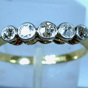 May include: A gold ring featuring five round, clear, faceted stones set in a horizontal row. The stones are held in place by silver-toned metal settings, creating a contrast against the thin, polished yellow gold band of the ring.