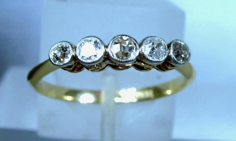 May include: A gold ring featuring five round, clear, faceted stones set in a horizontal row. The stones are held in place by silver-toned metal settings, creating a contrast against the thin, polished yellow gold band of the ring.