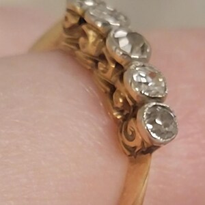May include: A gold ring featuring a row of five round, clear, faceted stones set in silver-toned metal settings. The band is made of yellow gold and displays a decorative, intricate gallery design beneath the stone settings.