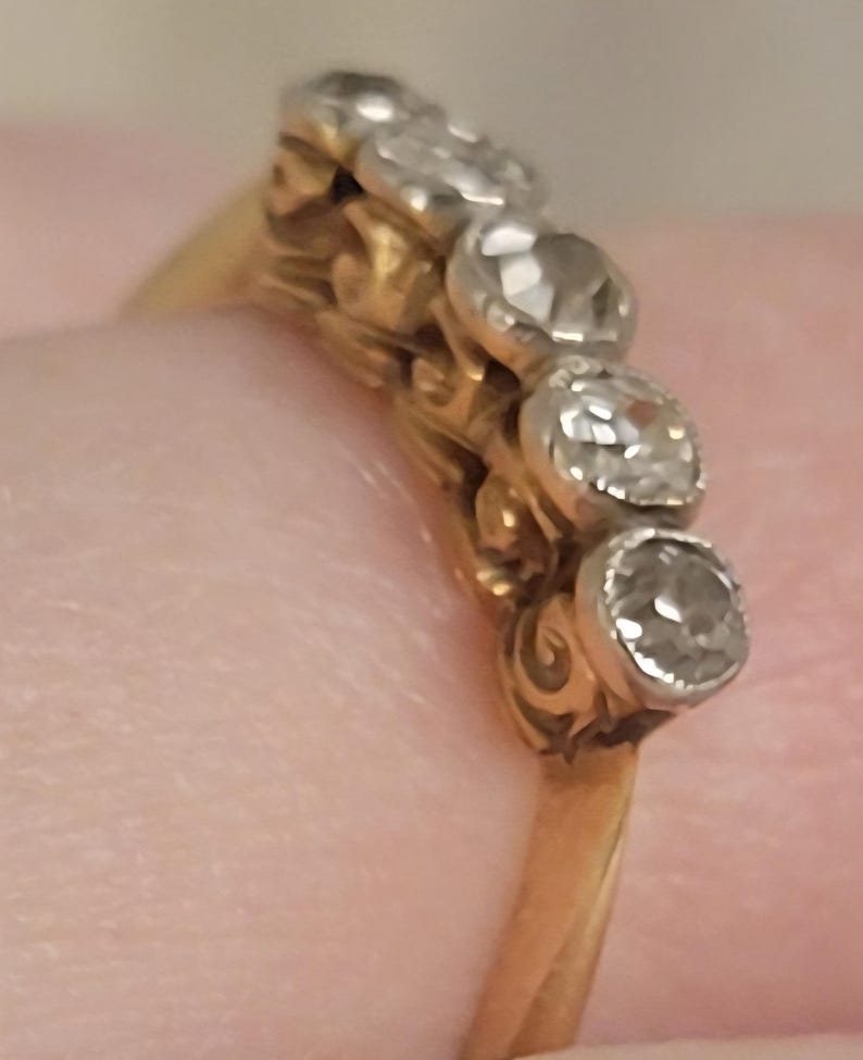 May include: A gold ring featuring a row of five round, clear, faceted stones set in silver-toned metal settings. The band is made of yellow gold and displays a decorative, intricate gallery design beneath the stone settings.