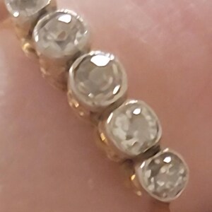 May include: A gold ring featuring five round, clear, faceted stones set in a row. Each stone is held in a circular metal setting, creating a continuous line of gems across the top of the band. The metal band appears to be yellow gold.