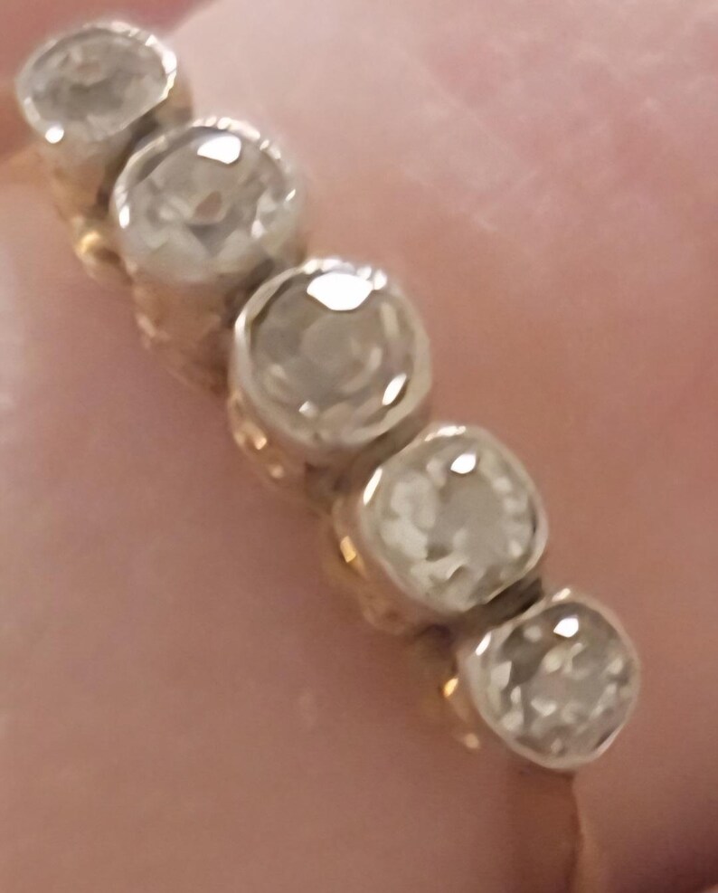 May include: A gold ring featuring five round, clear, faceted stones set in a row. Each stone is held in a circular metal setting, creating a continuous line of gems across the top of the band. The metal band appears to be yellow gold.