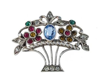 May include: A silver brooch featuring a basket design, embellished with colourful gemstones. The basket is filled with flowers crafted from pink, red, yellow, and green stones. A blue gemstone sits at the centre of the basket.