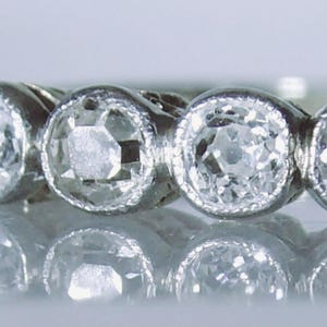 May include: A silver-toned ring featuring five round, faceted clear stones set in a row. Each stone is encased in a circular bezel setting with a fine milgrain edge detail. The band is metallic and reflects light, showcasing a classic design.