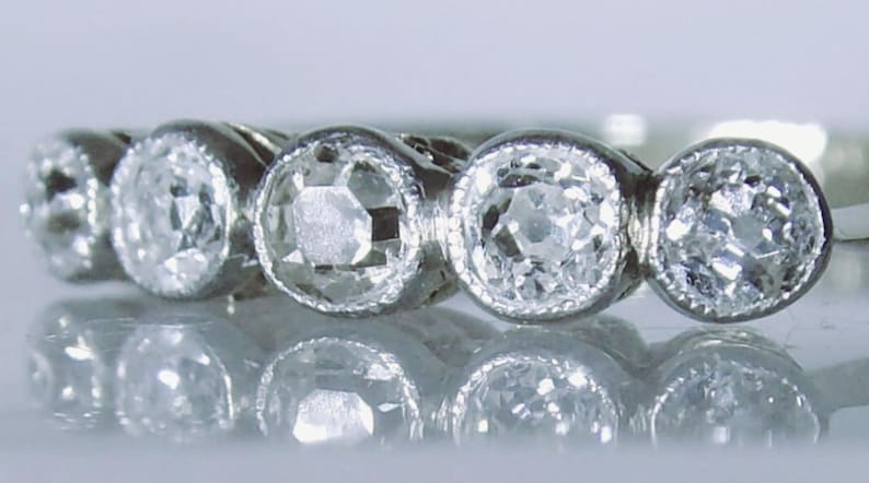 May include: A silver-toned ring featuring five round, faceted clear stones set in a row. Each stone is encased in a circular bezel setting with a fine milgrain edge detail. The band is metallic and reflects light, showcasing a classic design.