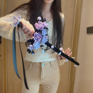 Puede incluir: Una empuñadura de espada decorativa envuelta en tela negra, adornada con racimos de flores de tela de color morado claro y hojas oscuras. Una cinta negra larga cuelga de la guarda y pequeños detalles blancos están fijados a lo largo del mango.