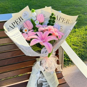 May include: A bouquet of pink and white flowers wrapped in newspaper. The bouquet includes pink lilies, tulips, and small white flowers. The bouquet is tied with a pink ribbon and rests on a wooden bench.