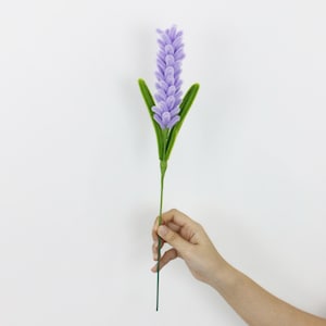 May include: A single artificial lavender stem featuring a tall, textured purple flower spike composed of small, rounded segments. The stem includes several pointed, vibrant green leaves extending from the base of the purple floral section.