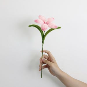 May include: A single artificial flower with five light pink petals and a central cluster of white stamens. The flower is attached to a thin green stem with two curved, fuzzy green leaves extending outward from the base of the pink blossom.