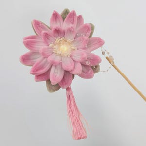 May include: A pink fabric flower hair accessory featuring layered petals with a central bead, green fabric leaves, a dangling pink thread tassel, and a thin wooden stick attachment adorned with a delicate string of small clear beads.