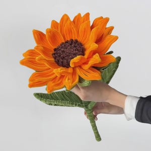 Può includere: Un girasole di peluche con petali in velluto arancione brillante e un centro testurizzato marrone scuro. Il fiore presenta un lungo stelo verde con una singola foglia verde attaccata, realizzato in un tessuto morbido simile al velluto a coste.