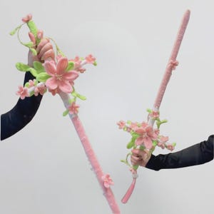 Puede incluir: Dos espadas de peluche con una hoja de color rosa suave y un mango decorado con enredaderas de tela verde claro y múltiples flores de cerezo rosas. Las espadas tienen una apariencia texturizada cubierta de tela con detalles florales.