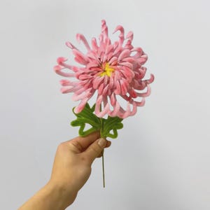 May include: A pink artificial chrysanthemum flower with long, curled petals and a yellow centre. The flower features a green stem with two green leaves attached. The petals have a soft, textured appearance, and the stem is thin and straight.
