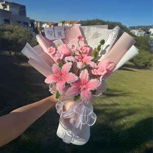 Op de afbeelding: Een boeket van roze stoffen bloemen met wit en roze papieren verpakking. Het boeket heeft grote, stervormige bloemen en kleinere bloesems, geaccentueerd met wit kant en parels. Het boeket wordt in een witte vaas gehouden.