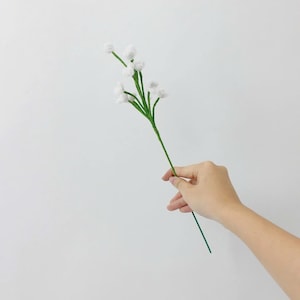 May include: A single artificial floral stem featuring a thin green wire stalk that branches into several smaller stems, each topped with a small, round, fuzzy white pom-pom blossom resembling a sprig of baby's breath against a plain background.