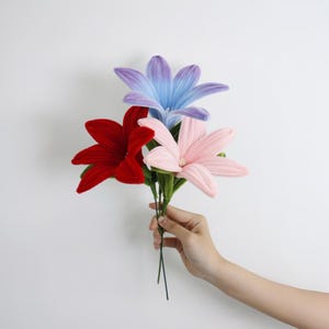 May include: A bouquet of three artificial flowers featuring a deep red lily, a soft pastel pink lily, and a vibrant blue and purple gradient lily. Each flower has six petals with a textured surface, green stems, and small white central stamens.