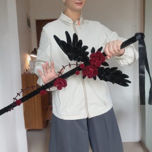 May include: A long, slender staff wrapped in black velvet, adorned with dark red fabric roses and black velvet wing-like attachments. The staff features a thin, winding vine-like element with small red floral accents and a black satin ribbon at the end.