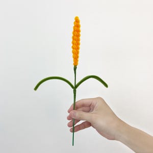 May include: A craft stem made from fuzzy pipe cleaners featuring a bright yellow, spiraled, cylindrical top section resembling a wheat stalk or flower head, attached to a thin green stem with two curved green leaves extending from the center.