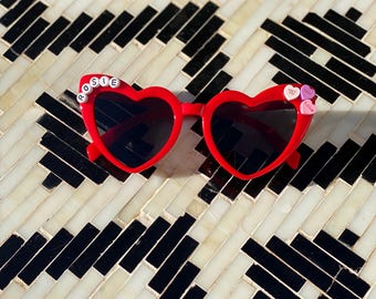 May include: Red heart-shaped sunglasses with black lenses and the name "ROSIE" spelled out in white letters on the left frame. The right frame has three small pink and white candy hearts with the words "Love", "You" and "Forever" on them.