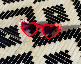 May include: Red heart-shaped sunglasses with the word "WINNIE" spelled out in white beads on the frame. The sunglasses are decorated with small candy hearts.