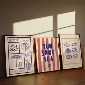 May include: Three framed prints with a beach theme. The left print features a blue and white illustration of a beach umbrella, a beach chair, a wave, and a person wearing a hat. The middle print has red and white stripes with the text "SUN SAND SEA". The right print features a blue and white illustration of various seashells.